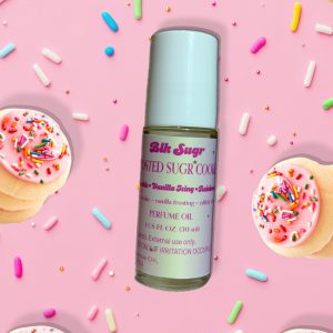 A bottle of Blk Sugr “Frosted Sugr Cookie” perfume oil on a pink background, surrounded by sugar cookies with pink icing and colorful sprinkles. The label includes notes of cookie, vanilla icing, and edible vibes.