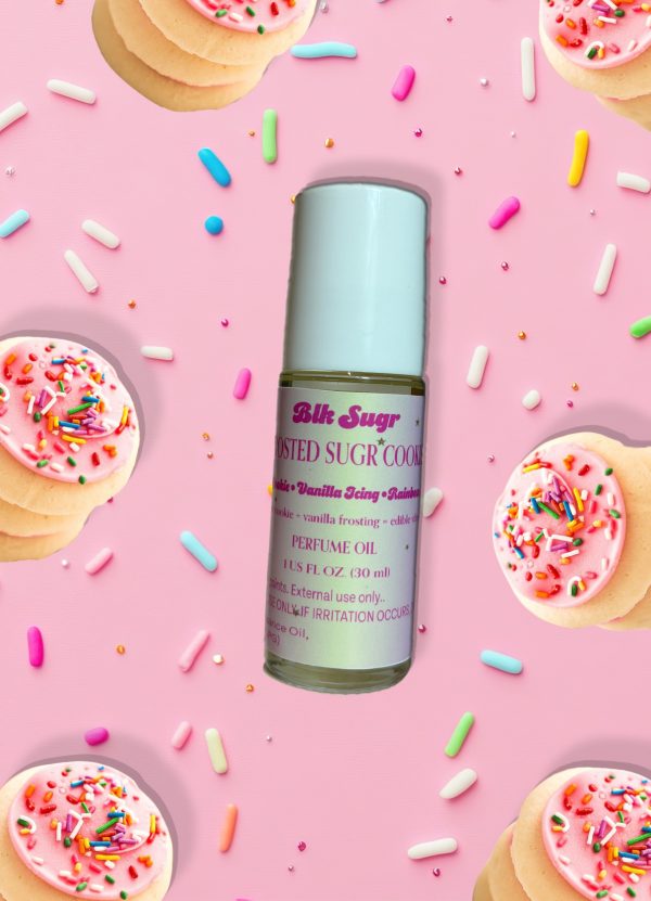 A bottle of Blk Sugr “Frosted Sugr Cookie” perfume oil on a pink background, surrounded by sugar cookies with pink icing and colorful sprinkles. The label includes notes of cookie, vanilla icing, and edible vibes.