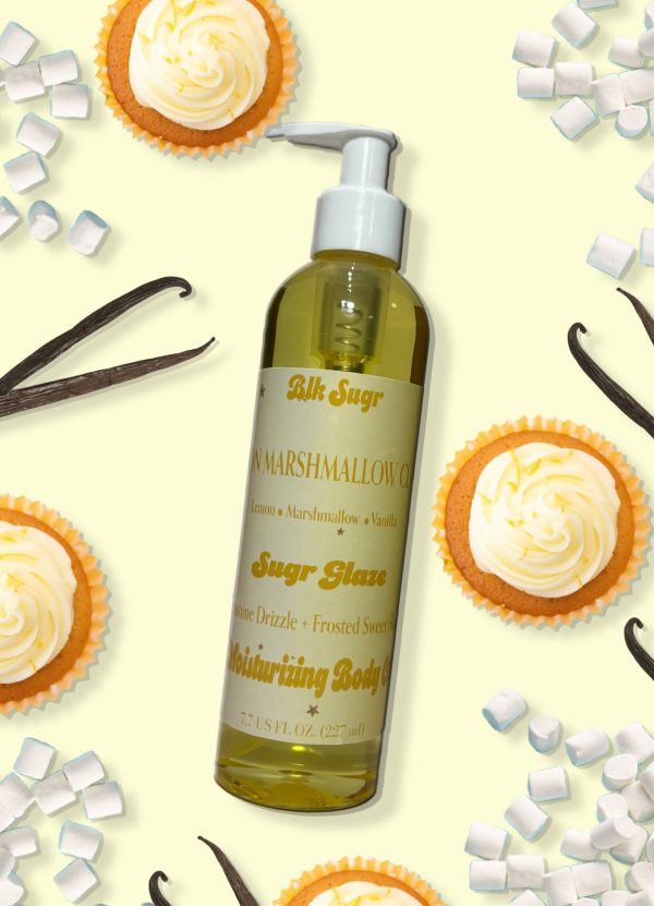 Blk Sugr “Sugr Glaze” moisturizing body oil bottle surrounded by lemon cupcakes, vanilla beans, and mini marshmallows on a soft yellow background. The label highlights notes of lemon, marshmallow, and vanilla.