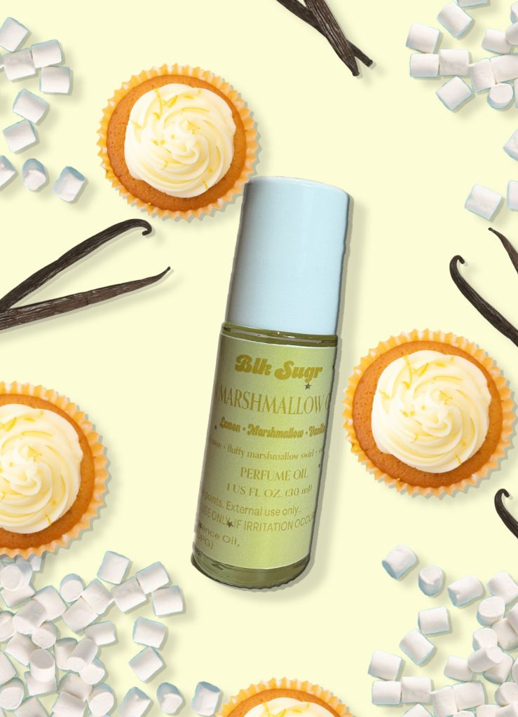 A bottle of Blk Sugr “Marshmallow” perfume oil on a pale yellow background, surrounded by lemon cupcakes, mini marshmallows, and vanilla bean pods. The label includes notes of lemon, marshmallow, and vanilla.