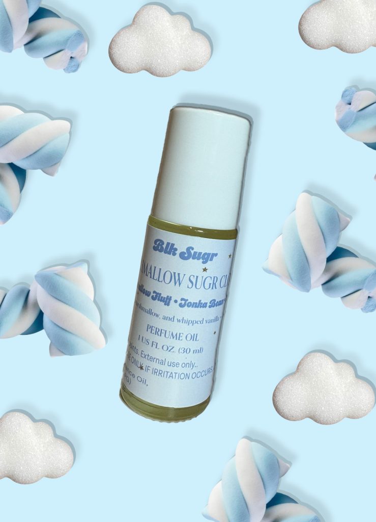 A bottle of Blk Sugr “Mallow Sugr Cloud” perfume oil on a baby blue background, surrounded by twisted marshmallows and fluffy white clouds. The label highlights marshmallow fluff, tonka bean, and whipped vanilla notes.