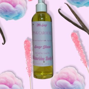 A bottle of Blk Sugr “Pink Carousel” Sugr Glaze body oil on a pastel pink background, surrounded by pink rock candy, cotton candy tufts, and vanilla beans. The label highlights notes of cotton candy, tonka bean, amber, and sugar crystals.