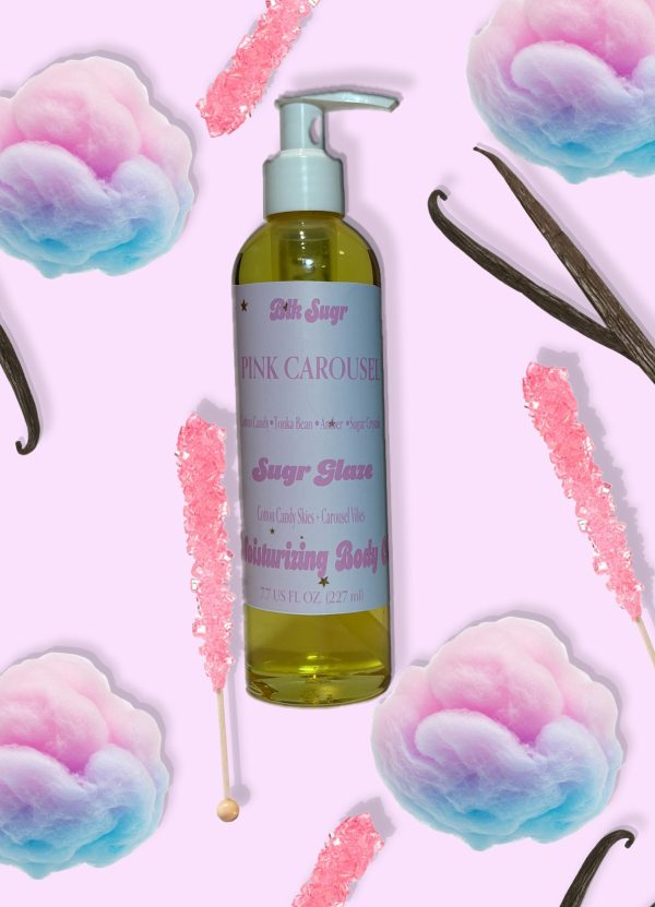 A bottle of Blk Sugr “Pink Carousel” Sugr Glaze body oil on a pastel pink background, surrounded by pink rock candy, cotton candy tufts, and vanilla beans. The label highlights notes of cotton candy, tonka bean, amber, and sugar crystals.