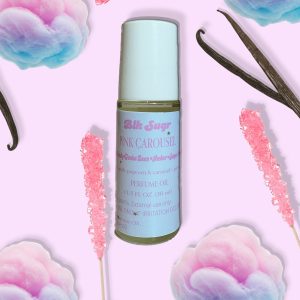 A bottle of Blk Sugr “Pink Carousel” perfume oil on a pink background, surrounded by pastel cotton candy, pink rock candy sticks, and vanilla beans. The label highlights notes of apple, popcorn, caramel, amber sugar, and tonka bean.