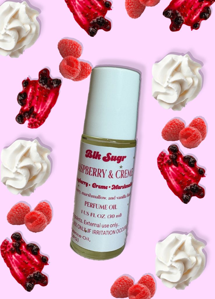 A bottle of Blk Sugr “Raspberry & Crème” perfume oil on a pastel pink background, surrounded by whipped cream, fresh raspberries, and raspberry drizzle. The label highlights notes of raspberry, crème, marshmallow, and vanilla fluff.
