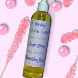 A bottle of Blk Sugr “Sugr Baby” Sugr Glaze body oil on a light pink background, surrounded by pink rock candy sticks and pink bubble gum balls. The label features notes of candied pear, caramel, and bubble gum.