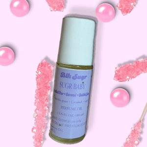 A bottle of Blk Sugr “Sugr Baby” perfume oil on a pastel pink background, surrounded by pink rock candy sticks and pink bubble gum balls. The label highlights bubble gum, caramel, and sugared pear notes.