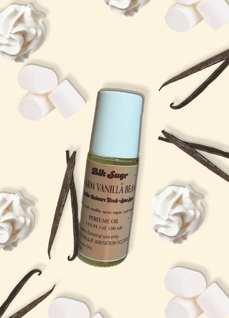 A bottle of Blk Sugr “Warm Vanilla Bean” perfume oil on a creamy beige background, surrounded by vanilla beans, whipped cream swirls, and marshmallows. The label notes include vanilla, cashmere woods, and spun sugar.