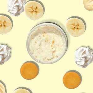 A jar of Blk Sugr Banana Pudding Sugr Buff surrounded by banana slices, vanilla wafers, whipped cream, and banana pudding swirls on a yellow background.