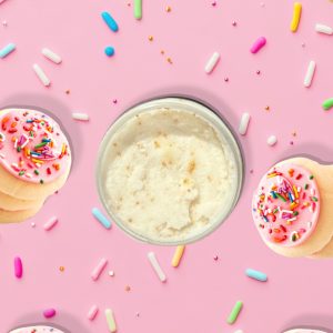 A jar of Blk Sugr Frosted Sugar Cookie Sugr Buff surrounded by pink frosted sugar cookies and rainbow sprinkles on a bright pink background.