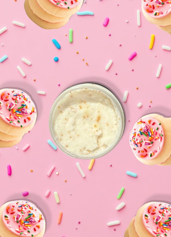 A jar of Blk Sugr Frosted Sugar Cookie Sugr Buff surrounded by pink frosted sugar cookies and rainbow sprinkles on a bright pink background.