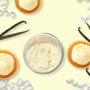 A jar of Blk Sugr Lemon Marshmallow Cupcake Sugr Buff surrounded by frosted lemon cupcakes, marshmallows, and vanilla bean pods on a light yellow background.
