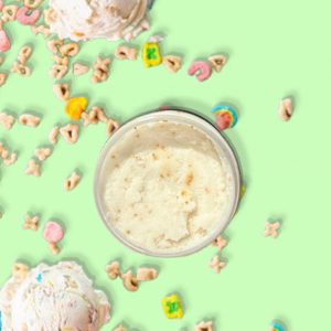 A jar of Blk Sugr Lucky Charms Ice Cream Sugr Buff surrounded by scoops of cereal ice cream and colorful cereal marshmallows on a mint green background.
