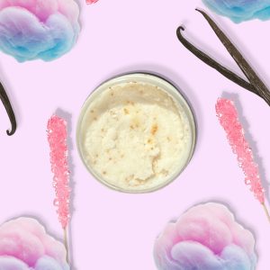 A jar of Blk Sugr Pink Carousel Sugr Buff surrounded by pastel cotton candy, vanilla beans, and pink rock candy sticks on a soft lilac background.