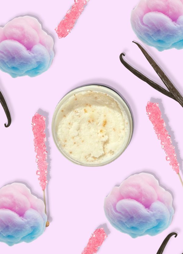 A jar of Blk Sugr Pink Carousel Sugr Buff surrounded by pastel cotton candy, vanilla beans, and pink rock candy sticks on a soft lilac background.