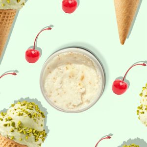A jar of Blk Sugr Pistachio Gelato Sugr Buff surrounded by pistachio gelato cones and bright red cherries on a mint green background.