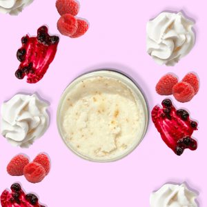 A jar of Blk Sugr Raspberry & Creme Sugr Buff surrounded by raspberries, raspberry jam swirls, and whipped cream on a pink background.