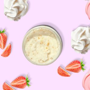A jar of Blk Sugr Strawberry Milk Sugr Buff surrounded by strawberries, whipped cream, and strawberry frosting swirls on a pink background.